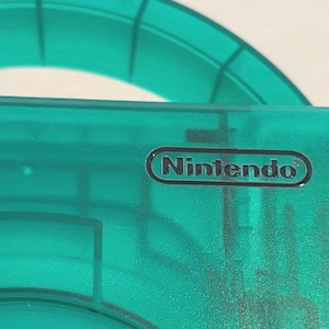May include: A translucent emerald green plastic gaming console shell featuring the Nintendo logo printed in black within a rounded rectangular border. The surface shows internal structural ribs and a circular cutout section visible through the material.