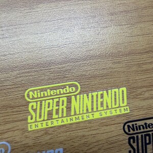 Op de afbeelding: Een close-up van een houten oppervlak met de gele tekst "Nintendo SUPER NINTENDO ENTERTAINMENT SYSTEM". Het Nintendo-logo staat boven de tekst. Andere Nintendo-logo's zijn zichtbaar.