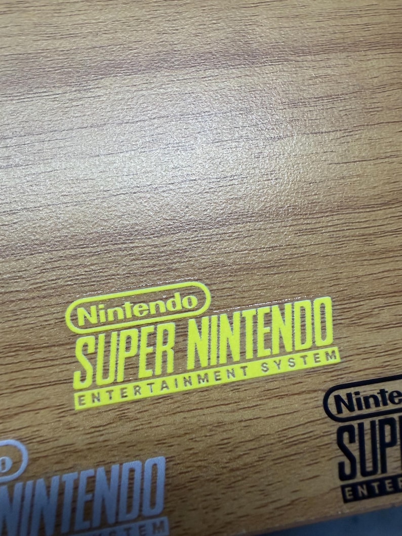 Op de afbeelding: Een close-up van een houten oppervlak met de gele tekst "Nintendo SUPER NINTENDO ENTERTAINMENT SYSTEM". Het Nintendo-logo staat boven de tekst. Andere Nintendo-logo's zijn zichtbaar.