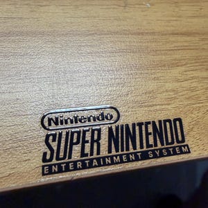 Op de afbeelding: Close-up van een houten oppervlak met het zwarte Nintendo Super Nintendo Entertainment System logo. Het logo is gecentreerd en toont de merknaam Nintendo boven de woorden "Super Nintendo".