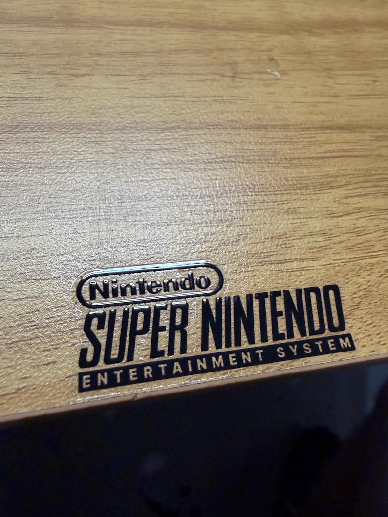 Op de afbeelding: Close-up van een houten oppervlak met het zwarte Nintendo Super Nintendo Entertainment System logo. Het logo is gecentreerd en toont de merknaam Nintendo boven de woorden "Super Nintendo".