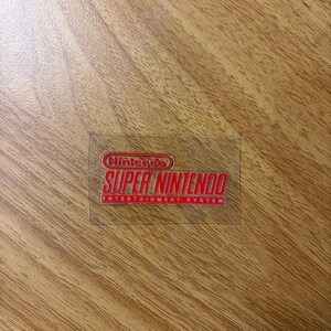 Op de afbeelding: Een transparante sticker met het rode Nintendo Super Nintendo Entertainment System logo. Het rechthoekige logo bevat het Nintendo-logo bovenaan, met "SUPER NINTENDO" eronder en "ENTERTAINMENT SYSTEM" onderaan.