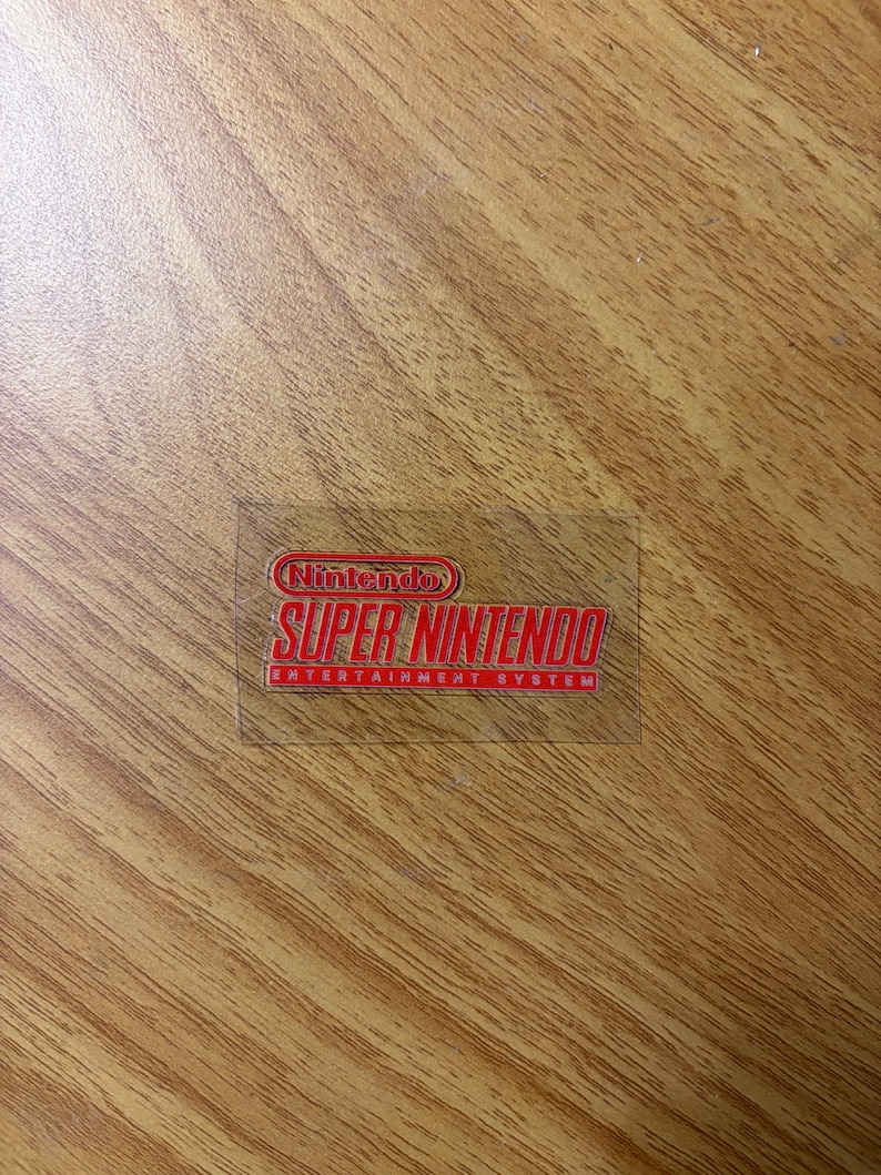 Op de afbeelding: Een transparante sticker met het rode Nintendo Super Nintendo Entertainment System logo. Het rechthoekige logo bevat het Nintendo-logo bovenaan, met "SUPER NINTENDO" eronder en "ENTERTAINMENT SYSTEM" onderaan.