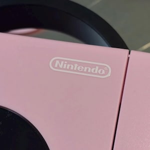 May include: A light pink electronic device featuring a black circular component and a black handle. The word Nintendo is printed in white text inside a white oval outline on the top surface of the pink casing, which has a smooth matte finish.