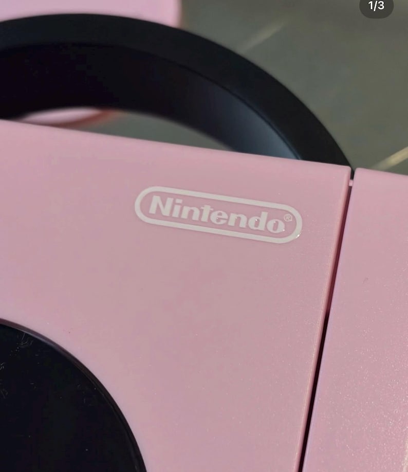 May include: A light pink electronic device featuring a black circular component and a black handle. The word Nintendo is printed in white text inside a white oval outline on the top surface of the pink casing, which has a smooth matte finish.