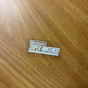 Op de afbeelding: Een doorzichtige sticker met het Nintendo-logo en de witte tekst "SUPER NINTENDO ENTERTAINMENT SYSTEM". De sticker is rechthoekig en ligt op een houten oppervlak.