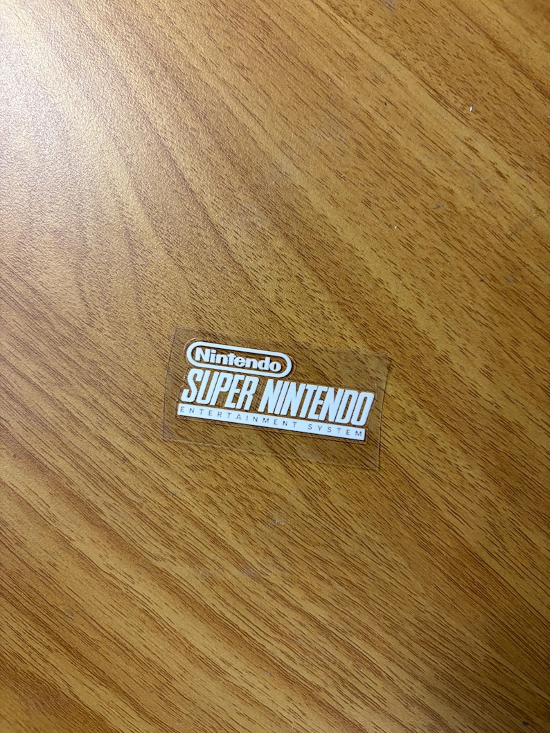 Op de afbeelding: Een doorzichtige sticker met het Nintendo-logo en de witte tekst "SUPER NINTENDO ENTERTAINMENT SYSTEM". De sticker is rechthoekig en ligt op een houten oppervlak.
