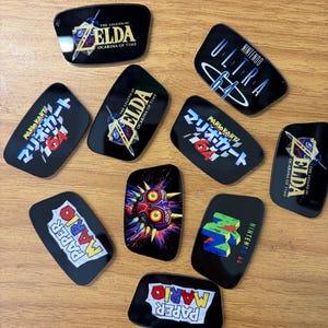 May include: A collection of ten rectangular black acrylic inserts featuring various Nintendo 64 game logos and artwork, including The Legend of Zelda: Ocarina of Time, Mario Kart 64, Paper Mario, Majora's Mask, and the Nintendo 64 logo in vibrant colours.