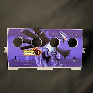 May include: A purple game console accessory featuring a graphic of a dragon-like creature and the text "Pokemon XD Gale of Darkness." The accessory has four circular holes and the text "SLOT-A" and "SLOT-B".