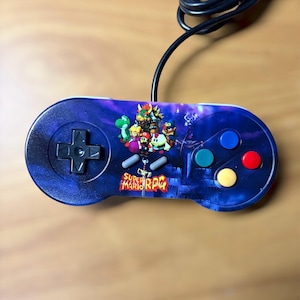 May include: A blue Super Nintendo-style controller with a colorful graphic featuring characters from Super Mario RPG. The controller has a directional pad, four colored action buttons, and two smaller buttons. A black cable extends from the top.