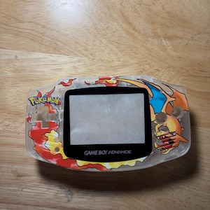 May include: A clear Game Boy Advance handheld gaming console featuring a custom graphic of the Pokemon Charizard with orange and yellow flames. The screen lens is black with the text GAME BOY ADVANCE printed in white at the bottom center.
