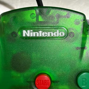 Puede incluir: Primer plano de un mando de Nintendo 64 verde translúcido. La palabra "Nintendo" está impresa en blanco. Se ve un botón "START" rojo, junto con un botón "B" verde. El cable del mando es negro.