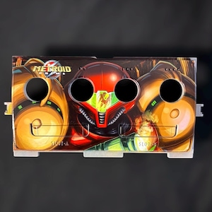 May include: A custom-designed game console faceplate featuring artwork from "Metroid Prime." The faceplate has a red, orange, and green color scheme, with the game's logo and character imagery. It has three circular openings and is labeled "SLOT A" and "SLOT B."