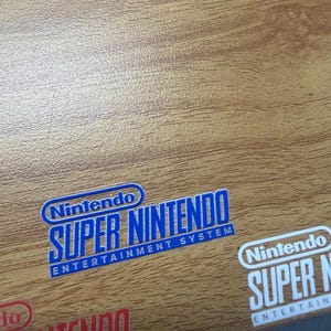 Op de afbeelding: Close-up van een houten oppervlak met meerdere stickers met het Nintendo Super Nintendo Entertainment System logo. De stickers zijn blauw, rood en wit, met de tekst in een vetgedrukte lettertype.