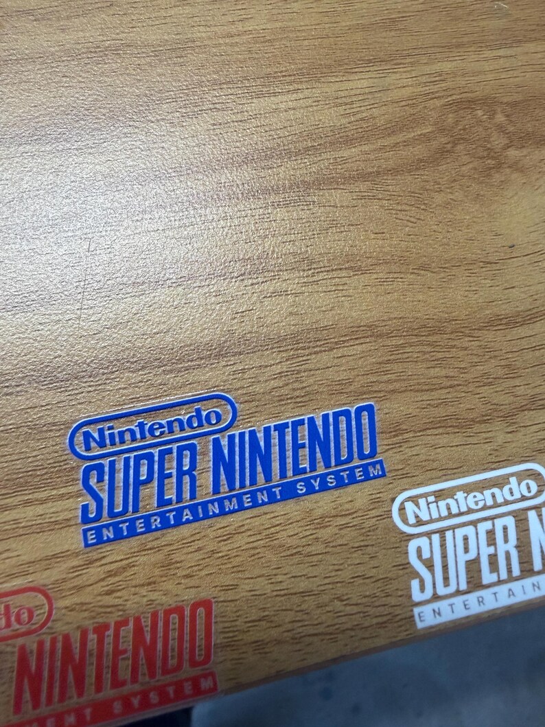 Op de afbeelding: Close-up van een houten oppervlak met meerdere stickers met het Nintendo Super Nintendo Entertainment System logo. De stickers zijn blauw, rood en wit, met de tekst in een vetgedrukte lettertype.