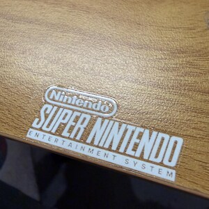 Op de afbeelding: Close-up van een houten oppervlak met een wit en zilver Nintendo Super Nintendo Entertainment System logo. Het logo is gecentreerd en bevat het Nintendo-logo boven de woorden "SUPER NINTENDO" en "ENTERTAINMENT SYSTEM".