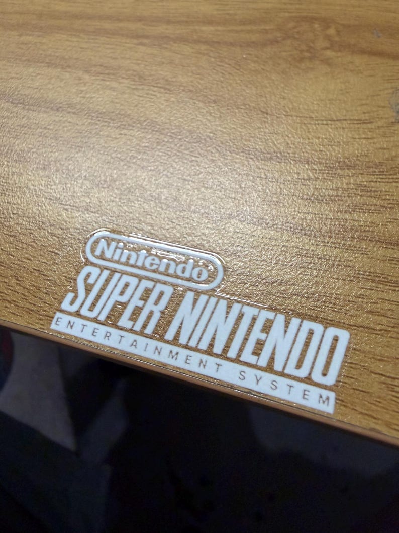 Op de afbeelding: Close-up van een houten oppervlak met een wit en zilver Nintendo Super Nintendo Entertainment System logo. Het logo is gecentreerd en bevat het Nintendo-logo boven de woorden "SUPER NINTENDO" en "ENTERTAINMENT SYSTEM".