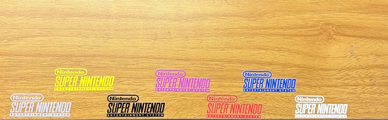 Op de afbeelding: Zeven vinylstickers met het logo van het Nintendo Super Nintendo Entertainment System in geel, roze, blauw, wit, zwart, rood en wit. Elke sticker toont de merknaam Nintendo boven de tekst Super Nintendo Entertainment System.