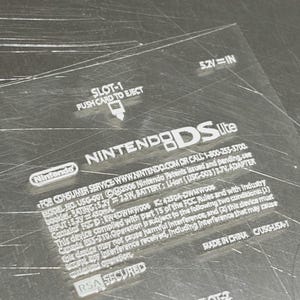 Può includere: Primo piano di una console Nintendo DS Lite argentata. La console presenta la scritta "Nintendo DS Lite". L'immagine mostra anche il testo "SLOT-1 PUSH CARD TO EJECT" e "SLOT-2".