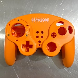 May include: An orange Nintendo GameCube Ultimate controller shell. The controller has a D-pad, analog stick, and button placements. The text "NINTENDO GAMECUBE ULTIMATE" is printed on the top of the controller.
