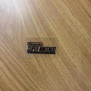 Op de afbeelding: Een transparante sticker met het Nintendo-logo en de tekst "SUPER NINTENDO ENTERTAINMENT SYSTEM" in zwart. De sticker bevindt zich op een houten oppervlak.