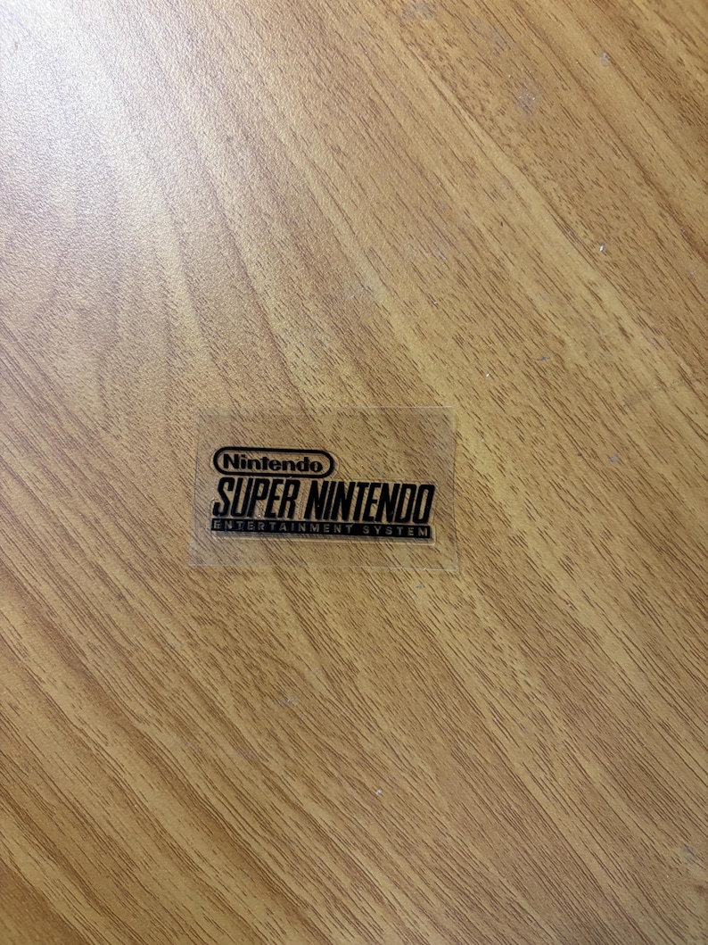 Op de afbeelding: Een transparante sticker met het Nintendo-logo en de tekst "SUPER NINTENDO ENTERTAINMENT SYSTEM" in zwart. De sticker bevindt zich op een houten oppervlak.