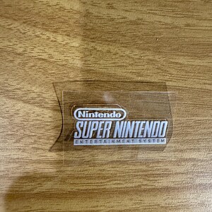 Op de afbeelding: Een transparante sticker met het Nintendo-logo en de tekst "SUPER NINTENDO ENTERTAINMENT SYSTEM" in het wit. De sticker is rechthoekig met afgeronde hoeken en ligt op een houten ondergrond.