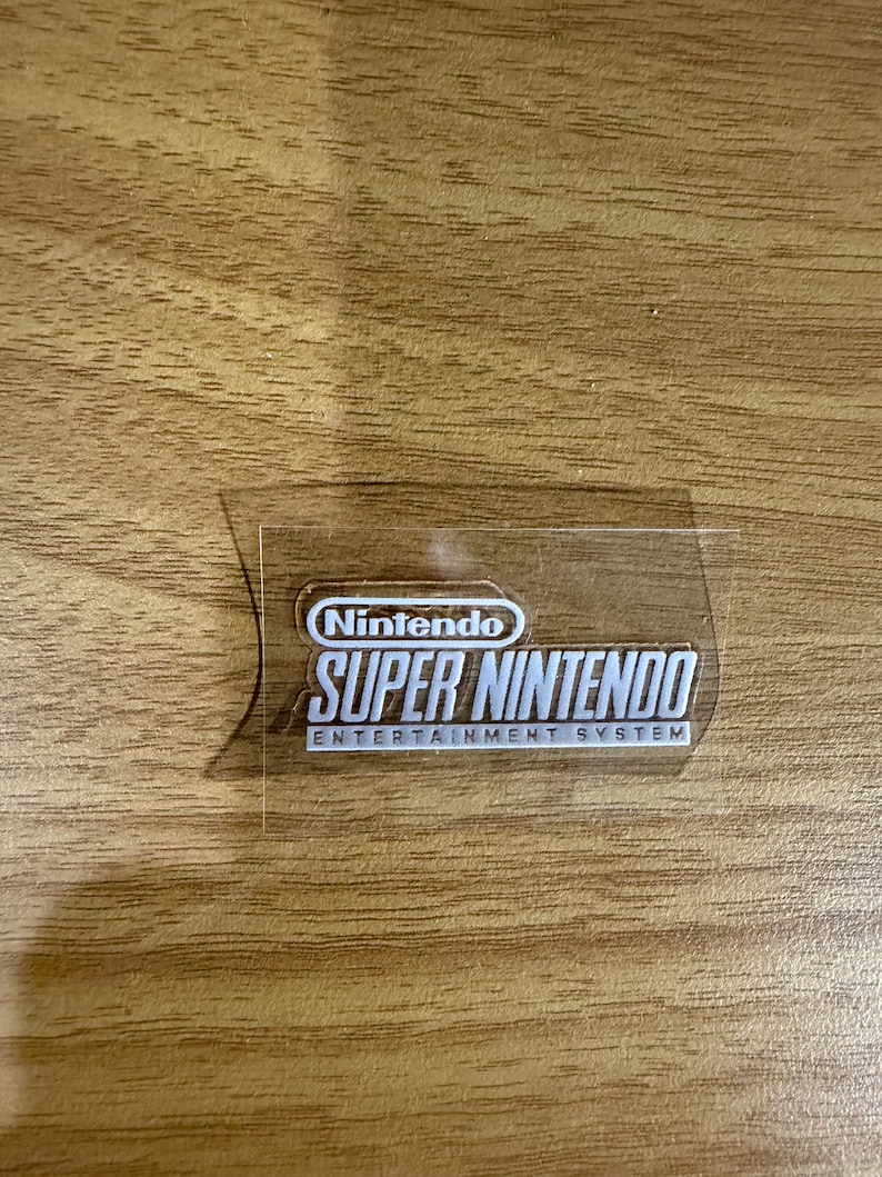 Op de afbeelding: Een transparante sticker met het Nintendo-logo en de tekst "SUPER NINTENDO ENTERTAINMENT SYSTEM" in het wit. De sticker is rechthoekig met afgeronde hoeken en ligt op een houten ondergrond.