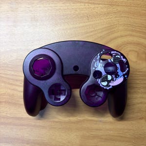 May include: A dark purple video game controller shell featuring a custom graphic on the right side. The graphic depicts a stylized black and white character with a long tongue and blue accents. The shell has circular cutouts for buttons.