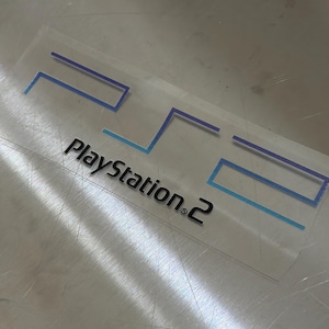 Może przedstawiać: Przezroczysta naklejka z logo PlayStation 2. Logo składa się z niebieskich i jasnoniebieskich linii, z czarnym napisem "PlayStation 2" poniżej. Naklejka znajduje się na metalowej powierzchni.