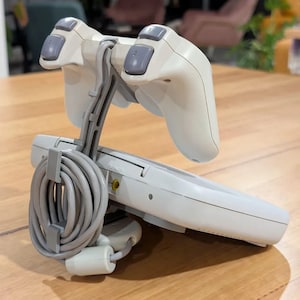 May include: A white and grey gaming console with two attached controllers. The controllers have grey buttons and are connected to the console by a grey cable. The console is resting on a wooden surface.