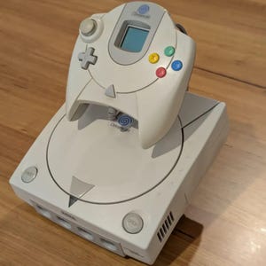 May include: A white Sega Dreamcast video game console with a matching controller. The console has a disc tray, power and open buttons, and the Dreamcast logo. The controller has a directional pad, analog stick, and colored action buttons.