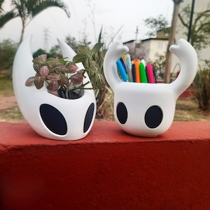 May include: Two white, stylized planters, one with a small plant with pink and green leaves, the other filled with colourful pens. Both have large, black oval eye-like shapes and a unique, curved design.