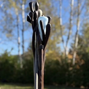 Könnte beinhalten: Eine hohe, braune Holzskulptur mit einem hellblauen, gebogenen Glaselement. Die Skulptur hat eine natürliche, organische Form mit mehreren Ästen und einer strukturierten Oberfläche. Der Hintergrund zeigt eine verschwommene Ansicht von Bäumen und blauem Himmel.