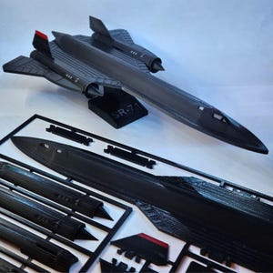 Puede incluir: Maqueta de avión SR-71 negra con detalles rojos en la cola. El modelo se exhibe sobre un soporte negro con la inscripción "SR-71". Se ven piezas sin montar en un marco negro, lo que sugiere un kit de modelo.
