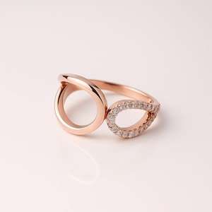 Könnte beinhalten: Roségoldener Ring mit einzigartigem Design. Eine Seite zeigt einen glatten, offenen Kreis, während die andere eine tropfenförmige Form mit kleinen, funkelnden Steinen aufweist. Der Ring ist vor weißem Hintergrund platziert.