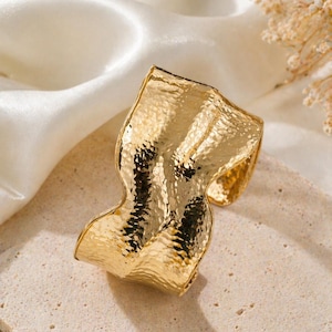 May include: A gold cuff bracelet with a textured, hammered finish. The bracelet has an undulating, irregular shape. It is displayed on a neutral surface with white satin fabric.