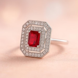 Könnte beinhalten: Silberner Ring mit einem rechteckigen, leuchtend roten Edelstein in der Mitte, umgeben von zwei Reihen kleiner, klarer Edelsteine. Der Ring hat ein klassisches, elegantes Design.