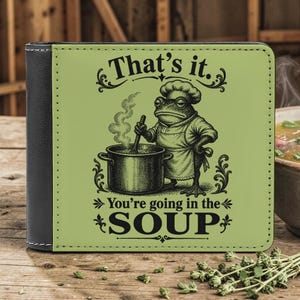 Könnte beinhalten: Ein schwarzes und grünes zweifach gefaltetes Portemonnaie mit einer Frosch-Koch-Illustration. Der Frosch rührt in einem Suppentopf, mit dem Text "That's it. You're going in the SOUP" darüber und darunter.