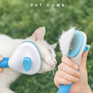 Peut inclure: Un peigne pour animaux de compagnie blanc et bleu brossant un chat blanc. Le peigne a une tête blanche avec un bouton bleu et une poignée bleue. Le texte "PET COMB" est en haut de l'image.