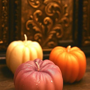 May include: Three pumpkin-shaped candles in shades of cream, mauve, and orange sit on a wooden surface. Each candle has a wick and a textured surface, evoking a fall or harvest theme. The background features ornate, golden wall decor.