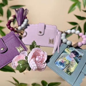 Puede incluir: Una colección de tarjeteros y llaveros de colores pastel. Los tarjeteros son morados, lavanda y verde azulado, con un emblema dorado. Un tarjetero verde azulado muestra una foto. También hay flores rosas decorativas y llaveros de cuentas.