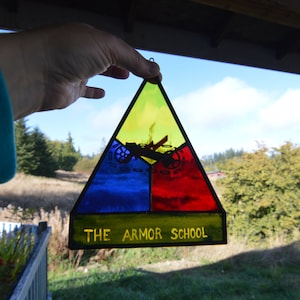 May include: A triangular stained glass window with a yellow, blue, and red design. The window features a black silhouette of a tank and the text "THE ARMOR SCHOOL".