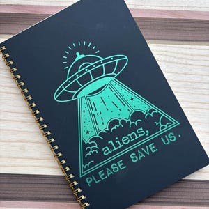 May include: A black spiral notebook with a green alien spaceship graphic and the text "aliens, please save us".