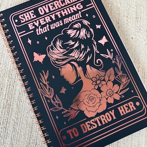 May include: A black spiral bound journal with a rose gold design. The cover features a woman's silhouette with flowers and butterflies. The text on the cover reads "She overcame everything that was meant to destroy her."
