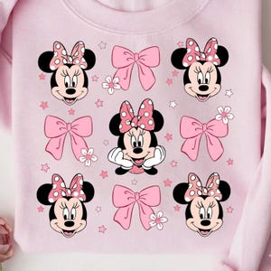 Op de afbeelding: Een lichtroze sweatshirt met een patroon van Minnie Mouse-gezichten, roze strikken, witte bloemen en roze sterren. Minnie Mouse heeft een stippenstrik en glimlacht. De sweatshirt heeft een ronde hals.
