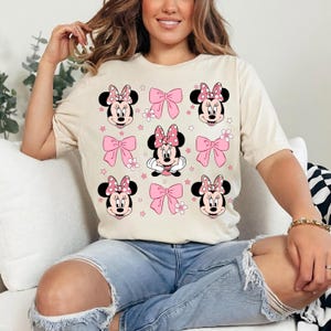 Op de afbeelding: Cr&egrave;mekleurig T-shirt met een herhaald patroon van Minnie Mouse-gezichten, roze strikken en bloemaccenten. Het ontwerp bevat Minnie's iconische polkadotstrik en lachende gezicht. Het shirt is een casual, comfortabele stijl.
