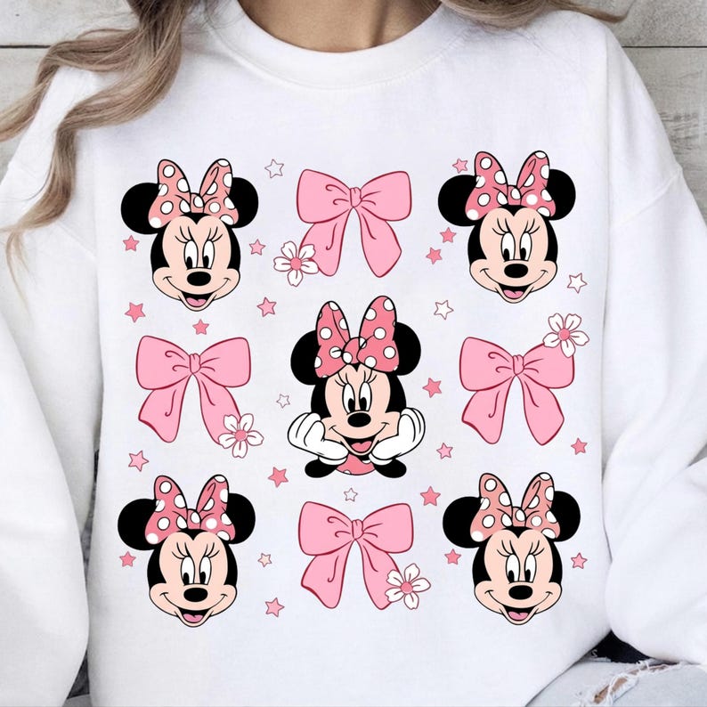 Op de afbeelding: Witte sweatshirt met een patroon van Minnie Mouse-gezichten, roze strikken, bloemen en sterren. Minnie Mouse heeft een roze strik met stippen. De strikken en sterren zijn roze. De sweatshirt is een casual, comfortabele stijl.