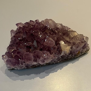 Könnte beinhalten: Ein Cluster aus Amethystkristallen, die einen tiefvioletten Farbton mit helleren, durchscheinenden Bereichen aufweisen. Die Kristalle sind dicht gepackt und bilden eine geodenartige Struktur. Die Basis ist ein rauer, natürlicher Stein. Die Gesamtform ist unregelmäßig und organisch.