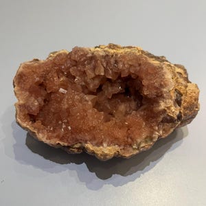 May include: A natural geode, featuring a cavity filled with light-pink crystals. The outer shell is a rough, earthy brown, contrasting with the delicate interior. The geode is displayed on a light grey surface.